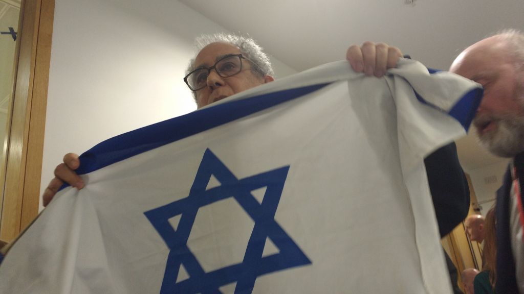 Pro-Israel activist Jonathan Hoffman being led out the room, holding an Israeli flag 