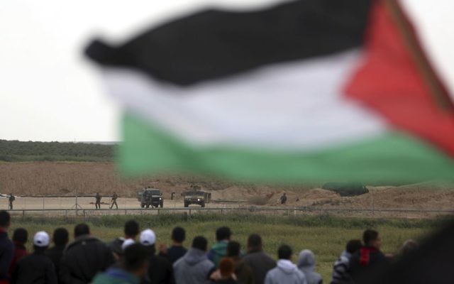 Protesters, one holding a Palestinian flag, stand in front of Israeli soldiers during a demonstration near the Gaza Strip border with Israel, in eastern Gaza City, Friday, March 30, 2018. (AP Photo/ Khalil Hamra)