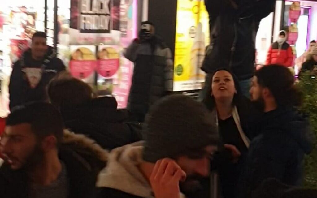 A Middle Eastern looking man in his 20s stands on a bench and makes insulting gestures towards the bus passengers.