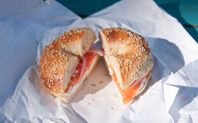 A sesame seed bagel with smoked salmon, cream cheese, onion and tomato, sitting on a paper wrapper.