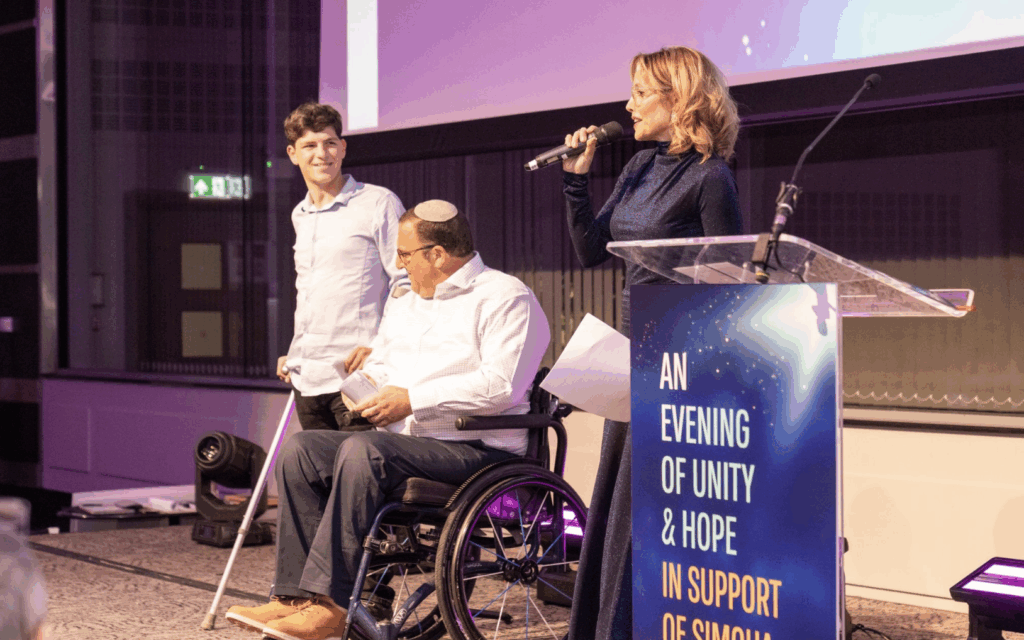 Itai and his father Ephraim share their story on stage as host Mili Avital addresses guests at Simcha LeYeled’s London fundraiser.